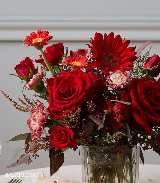 Vibrant red roses, gerberas, and carnations in clear glass vase with fresh foliage