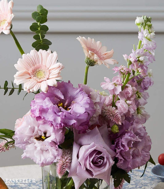 Fresh purple lisianthus and lavender roses with pink gerberas in clear glass vase, dining table arrangement
