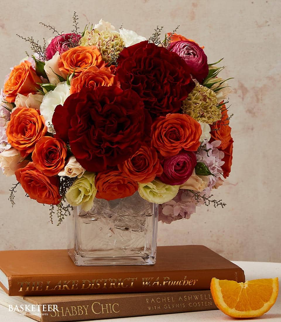 Vibrant amber and red rose arrangement in square glass vase with complementary orange and white blooms, styled on stacked