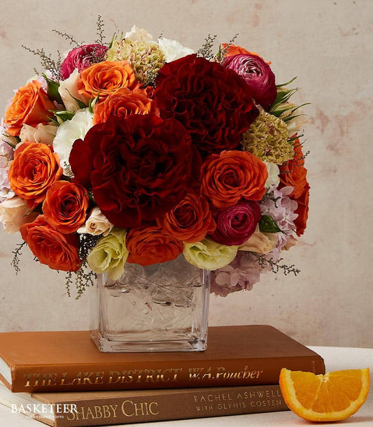 Vibrant amber and red rose arrangement in square glass vase with complementary orange and white blooms, styled on stacked