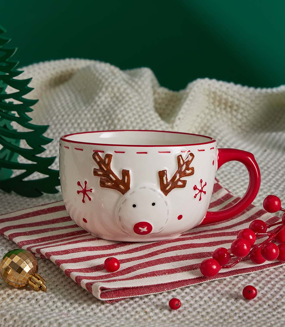 Reindeer ceramic mug with red berries and Christmas tree decoration on festive knitted blanket