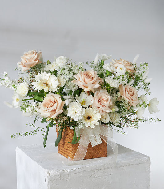 Cream roses and white gerberas in woven basket with trailing greenery and beige ribbon bow