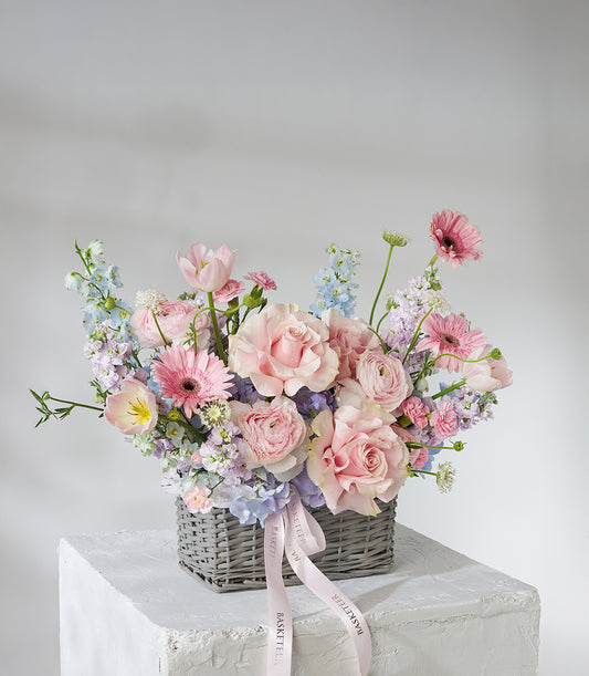 Pastel pink roses and gerbera daisies in soft blue and lavender blooms, arranged in grey woven basket with pink ribbon