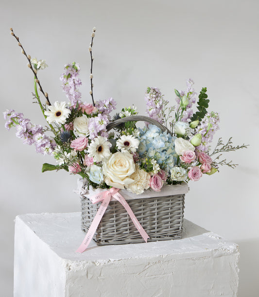 Pastel flower basket with white roses, blue hydrangeas, pink spray roses, and purple stock arranged in grey wicker
