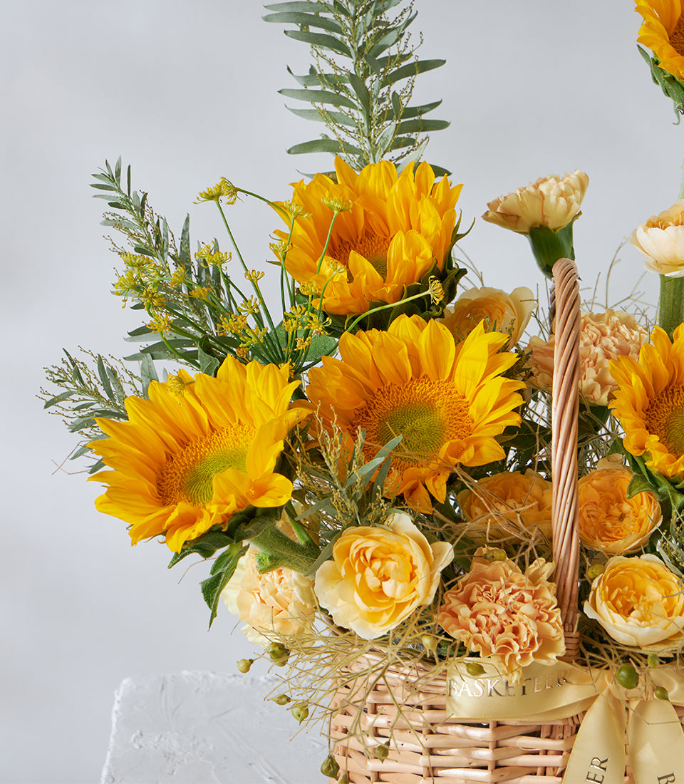 Vibrant sunflowers and yellow roses in a woven basket with peach carnations and green foliage