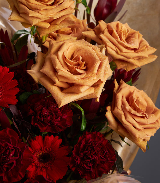 Warm peach roses and deep red gerberas with burgundy foliage in gold-wrapped luxury bouquet