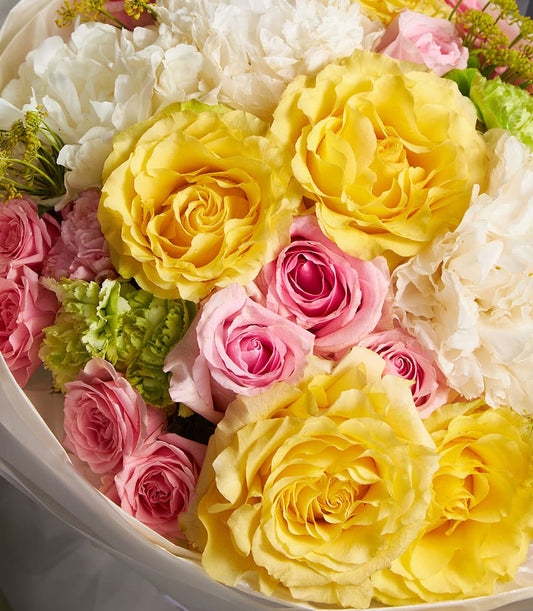 Vibrant mixed bouquet of golden yellow roses, pink roses, and white ruffled flowers with green accents, wrapped in clear