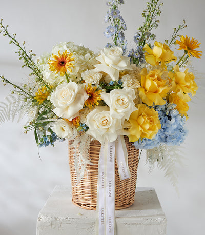 White and yellow roses with blue hydrangeas in woven wicker basket arrangement