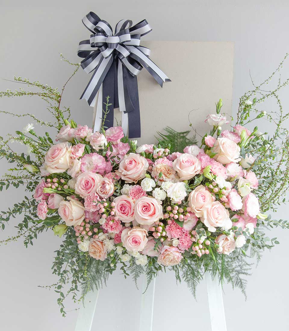 Blush rose and white flower wreath with greenery on easel stand, sympathy arrangement with striped ribbon bow