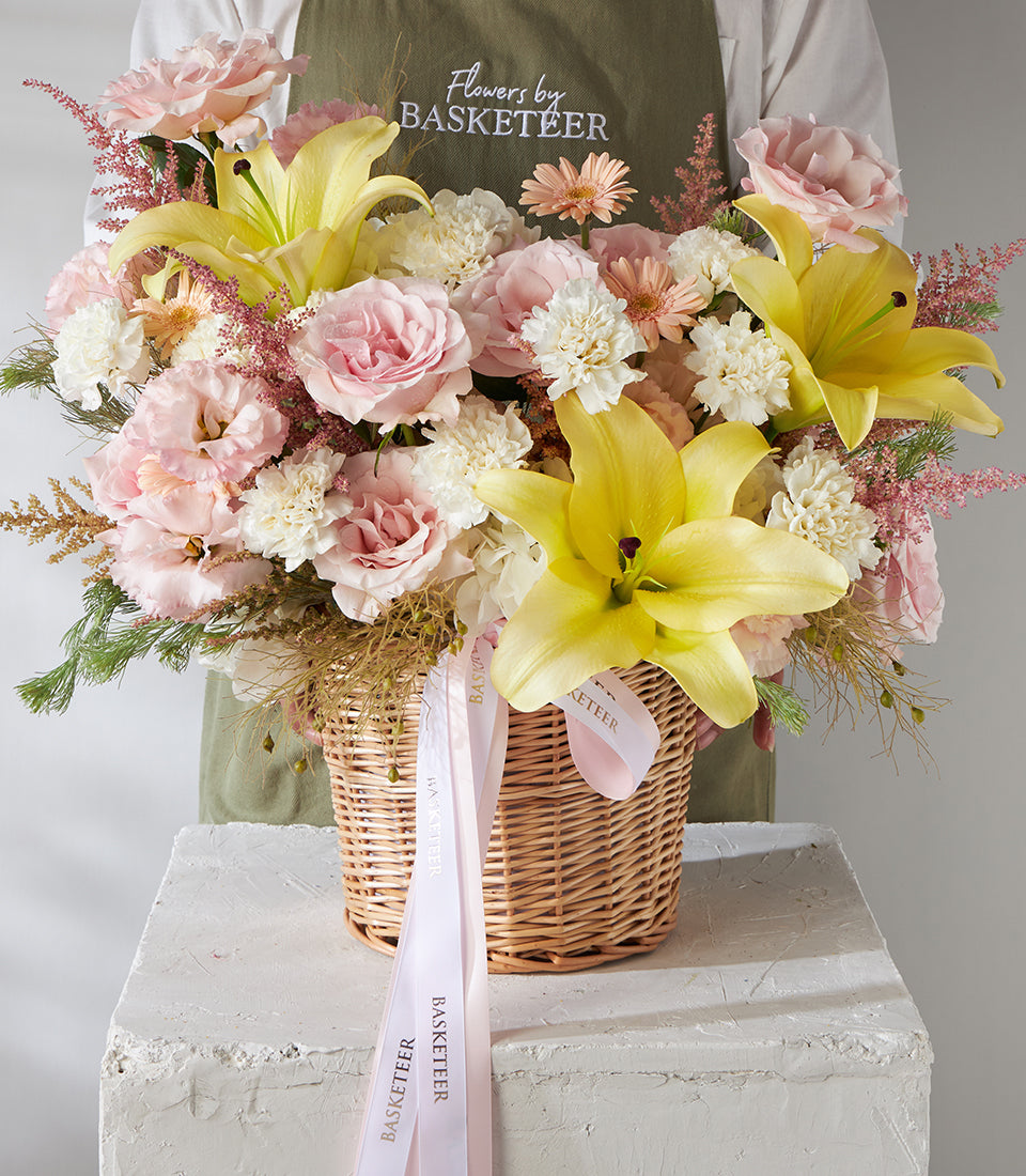 Vibrant yellow lilies and pink roses in woven basket with white carnations and green foliage