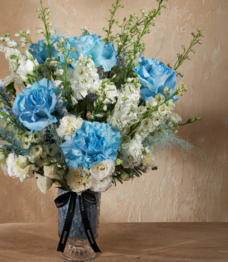 Light blue roses and ruffled carnations with cream florals in a clear glass vase, premium gift arrangement