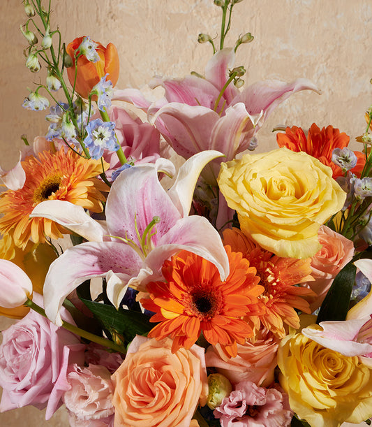 Radiant mix of pink lilies, orange gerberas, yellow roses, and delphiniums in glass vase arrangement