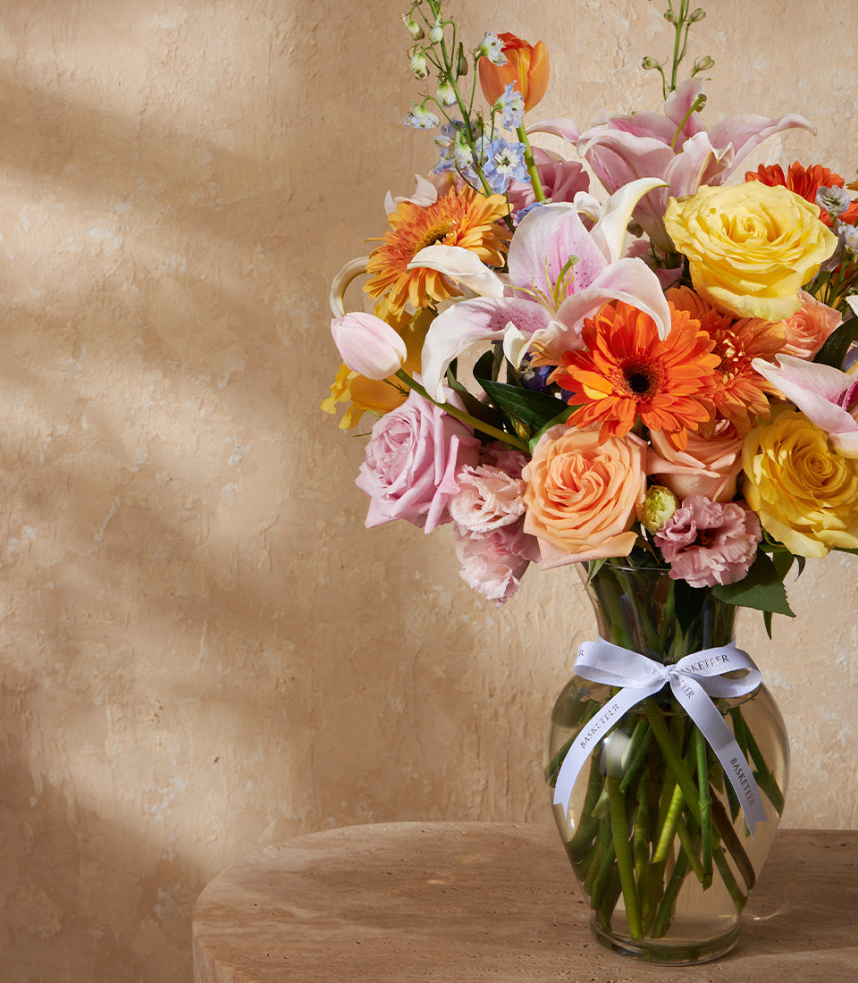 Vibrant mixed flower arrangement with pink roses, gerbera daisies, and lilies in clear glass vase with white ribbon