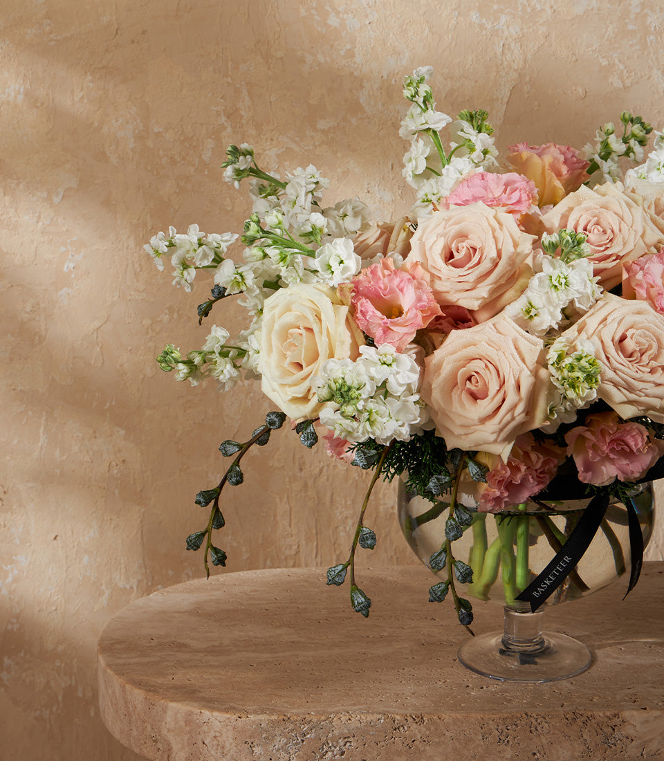 Elegant floral arrangement with pale pink roses, cream eustoma, and white stock flowers in clear glass footed vase