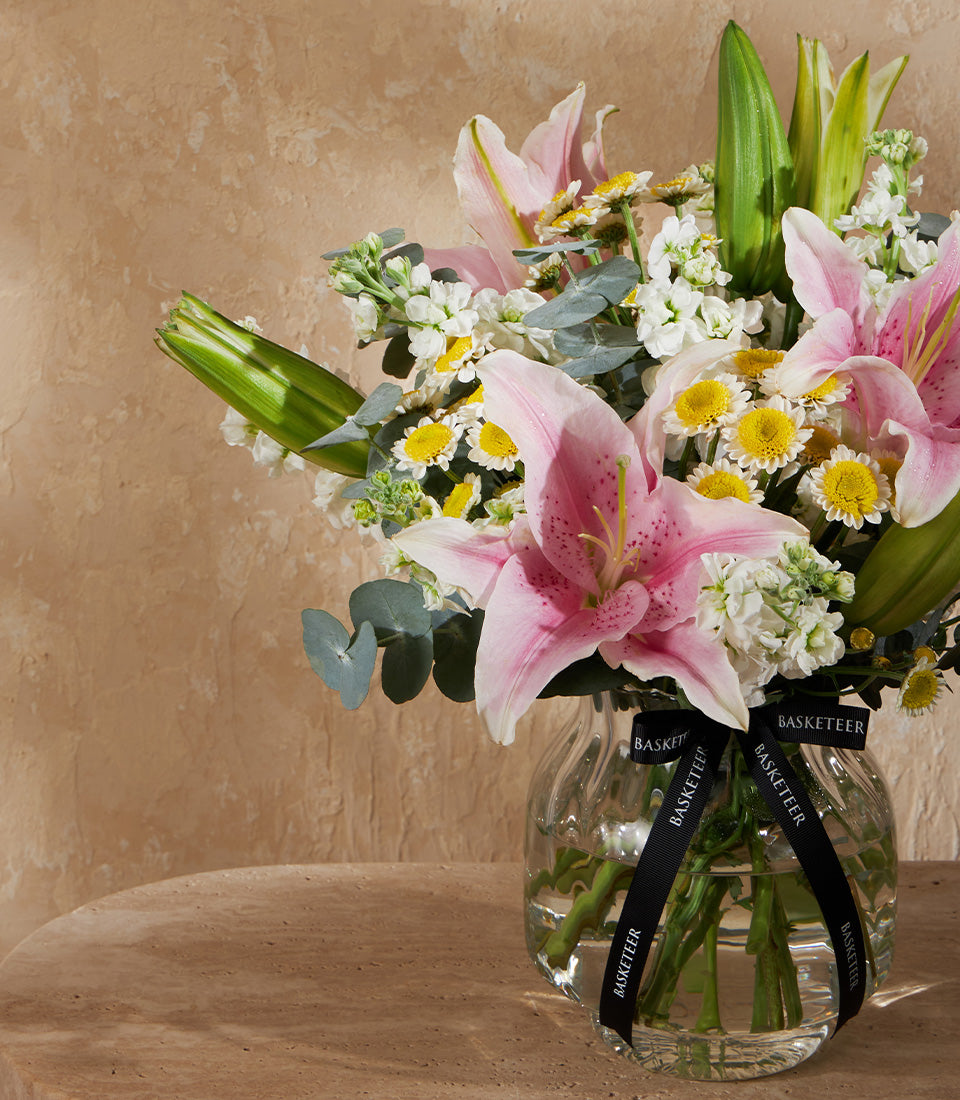 Pink stargazer lilies, white stock flowers, and yellow daisies in clear glass vase with Basketeer ribbon