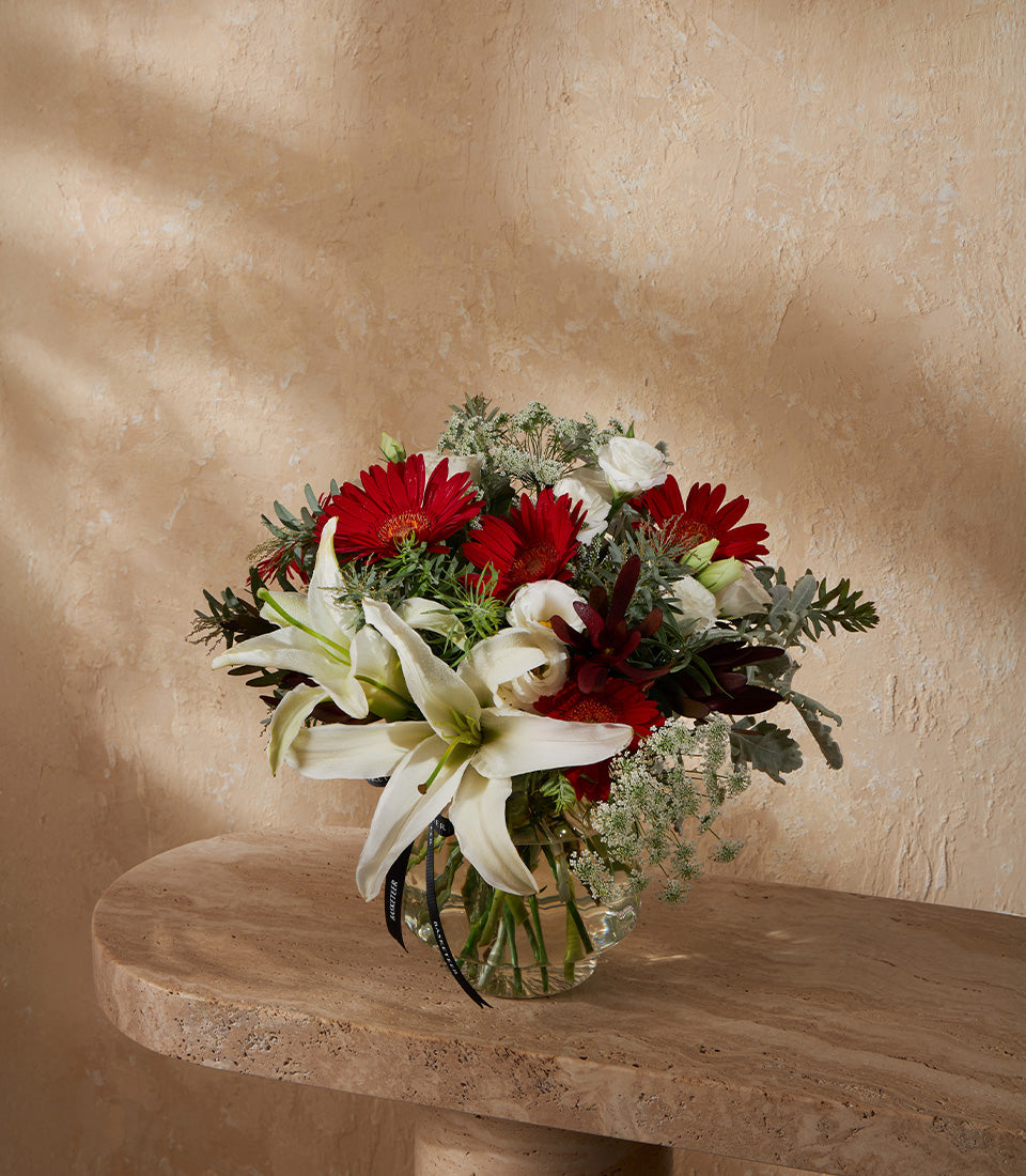 Vibrant red gerbera daisies and white lilies in clear glass vase with silk ribbon