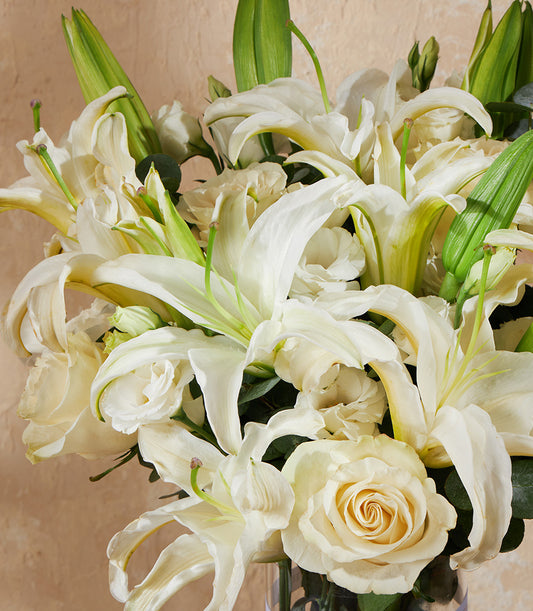 White lilies and roses with lush green foliage in clear glass vase arrangement