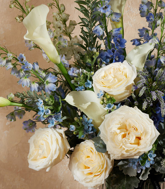 Lush white roses and calla lilies with blue delphiniums in premium golden vase arrangement