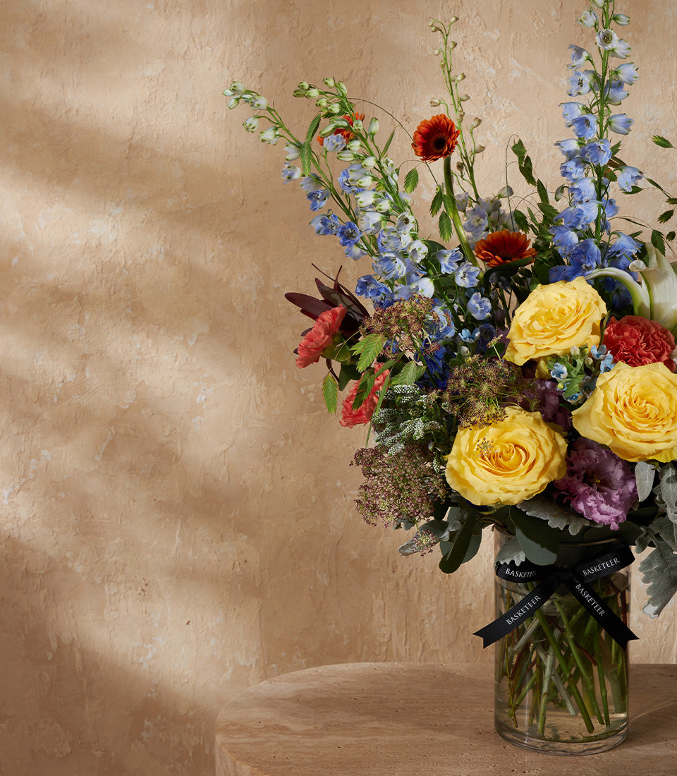 Vibrant yellow roses, blue delphiniums, and orange gerberas in clear glass vase with black ribbon