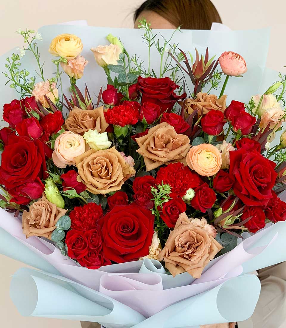 Luxe mixed rose bouquet in red, caramel, and peach with ranunculus, wrapped in soft blue and pink paper