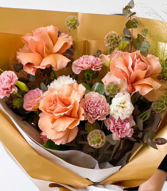 Peach and pink fresh rose bouquet with carnations and green foliage, wrapped in gold paper for graduation