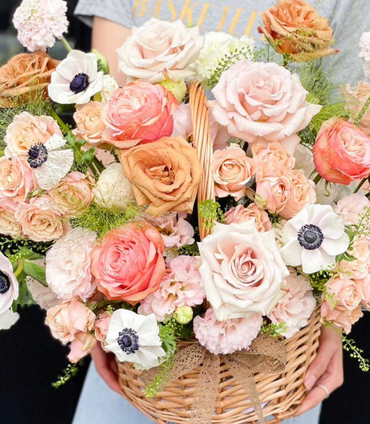 Lush peach and coral rose basket with white anemones, green foliage, and rustic brown ribbon