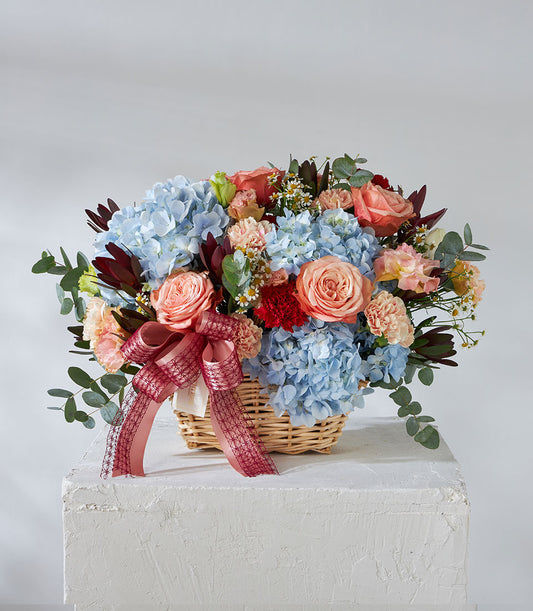 Vibrant get well flowers basket with blue hydrangeas, coral roses, and pink carnations in woven wicker