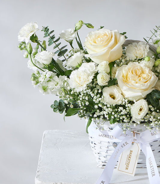 White and cream roses, lisianthus, and baby's breath in woven basket with eucalyptus and satin ribbon