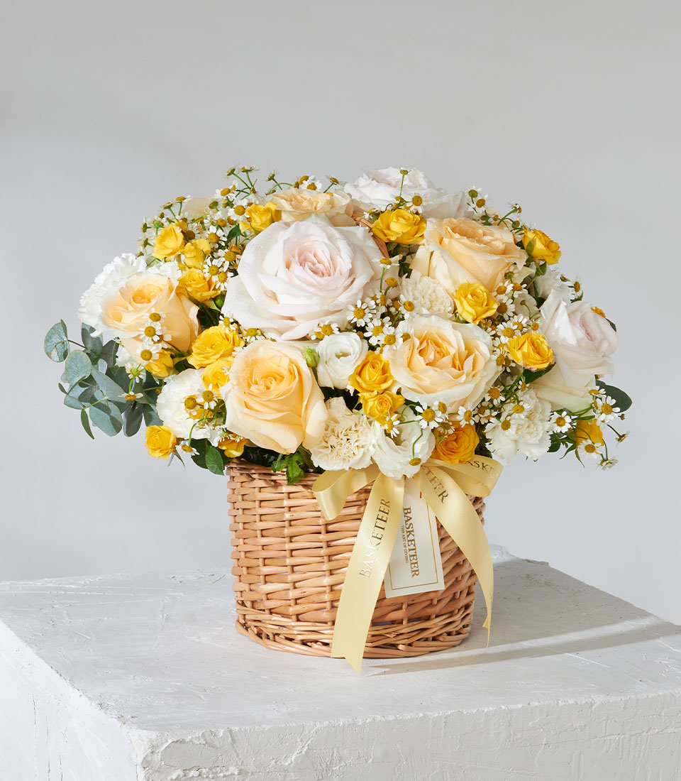 Pale yellow and blush rose arrangement in woven wicker basket with eucalyptus and white filler flowers