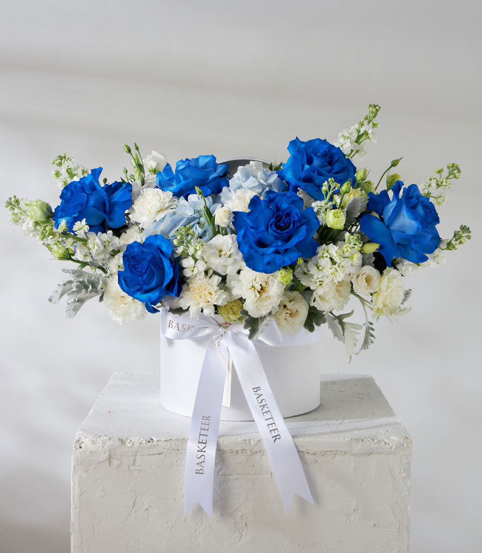 Tinted blue roses and white filler flowers in elegant white hatbox with satin ribbon