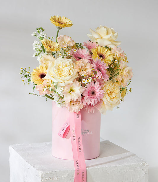 Pink velvet hatbox with roses, gerberas, and filler flowers in cream and yellow tones