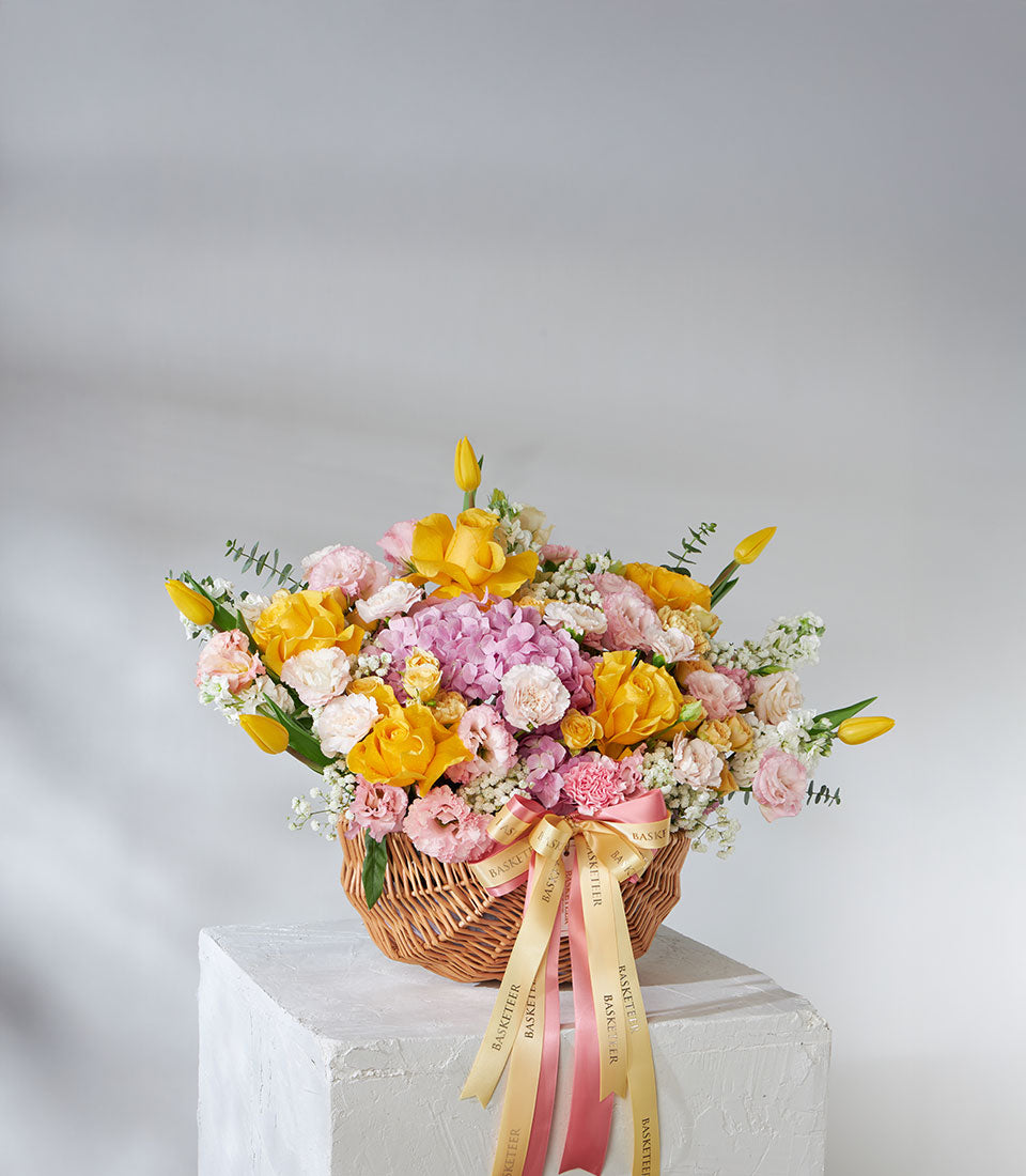 Vibrant yellow roses, pink hydrangeas, and purple flowers in woven wicker gift basket with satin ribbons