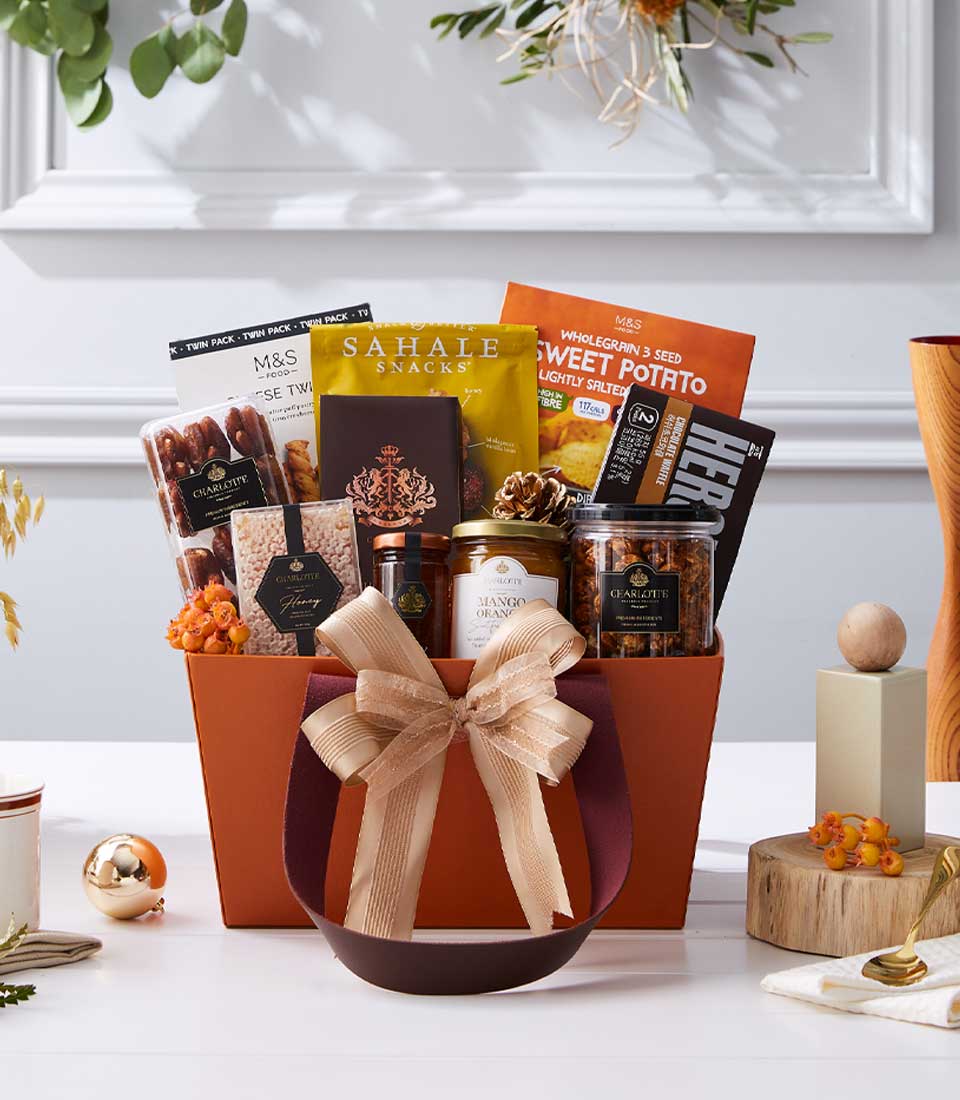 Luxury gourmet gift basket with chocolates, dates, and artisan snacks in deep orange-brown wicker with gold ribbon bow, New