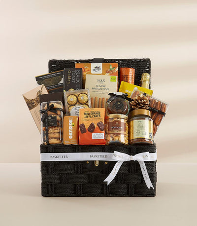 Elegant black wicker gourmet hamper with white ribbon bow, filled with premium snacks, chocolates, and sparkling beverage