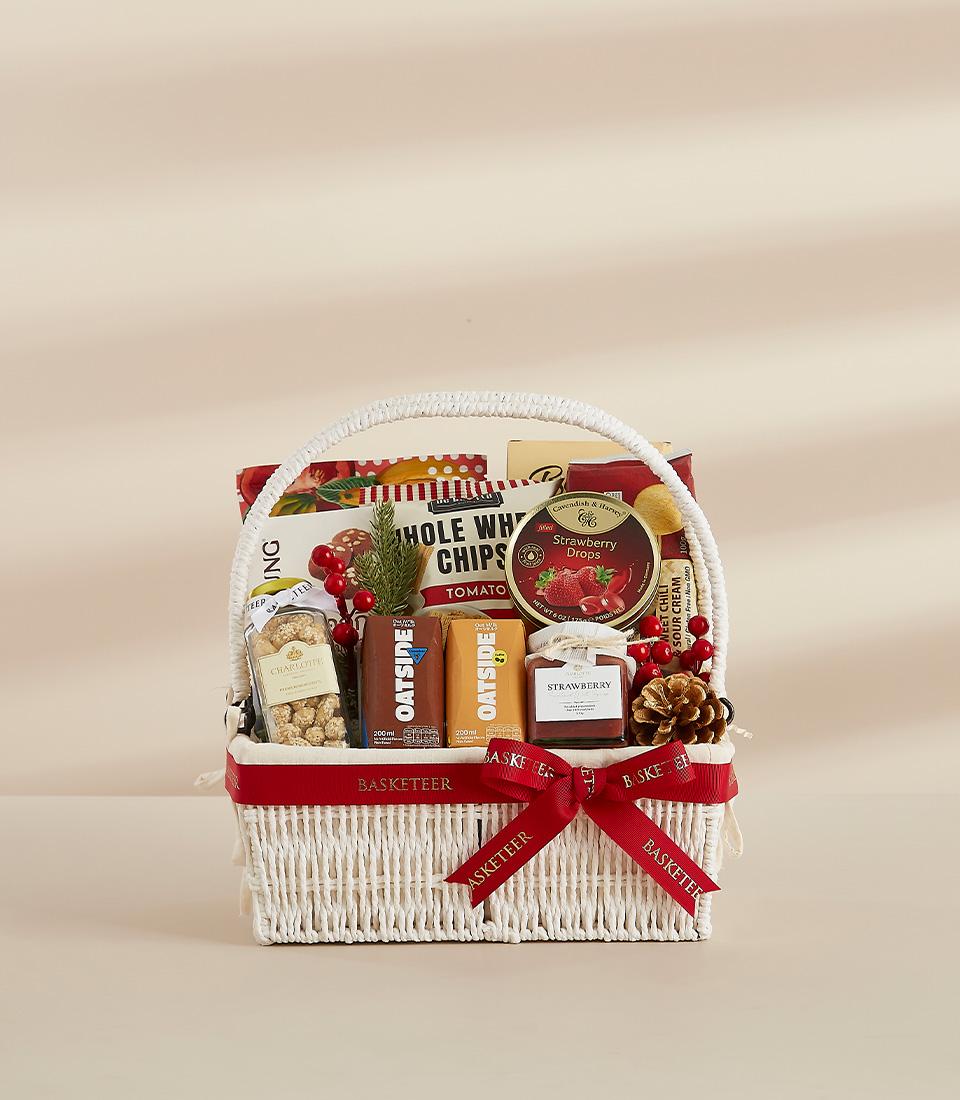 White woven gift basket filled with gourmet snacks, strawberry jam, and festive holiday treats tied with red bow
