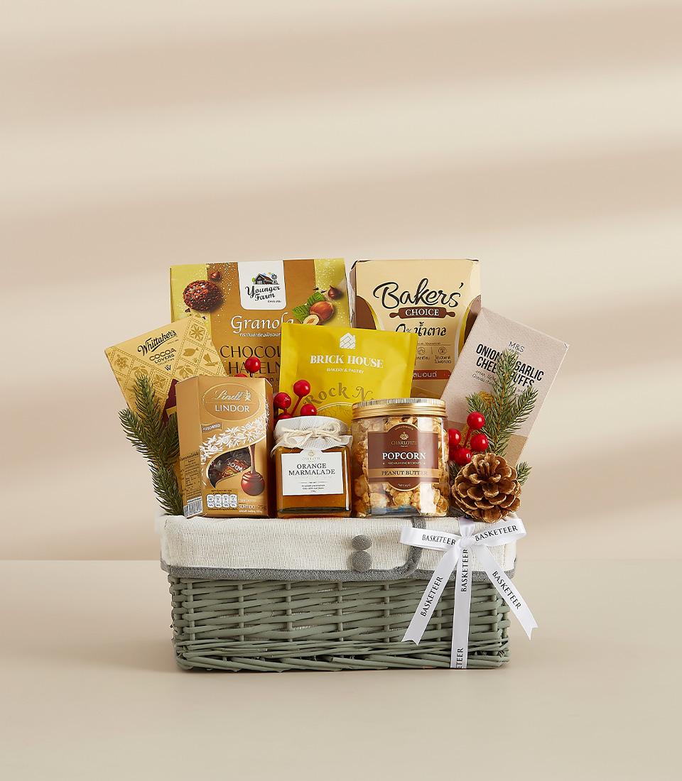 Woven grey gift basket filled with gourmet chocolates, cookies, marmalade, and gourmet treats, decorated with festive pine