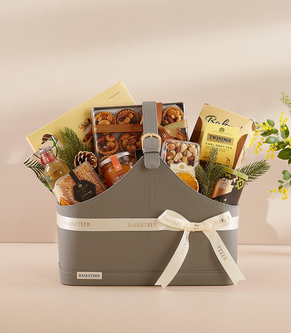 Grey faux leather gourmet gift bag with gold buckle, filled with premium teas, nuts, jam, and festive greenery for New Year