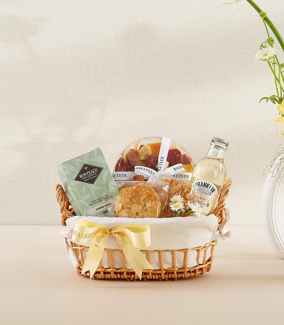 Woven wicker gift basket with jasmine green tea, dried fruits, ginger beverage, and almond florentine cookies, tied with