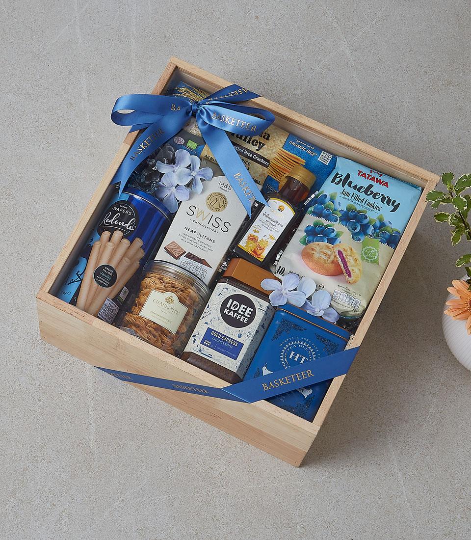 Wooden gift box filled with gourmet snacks, coffee, chocolate, and treats tied with blue ribbon