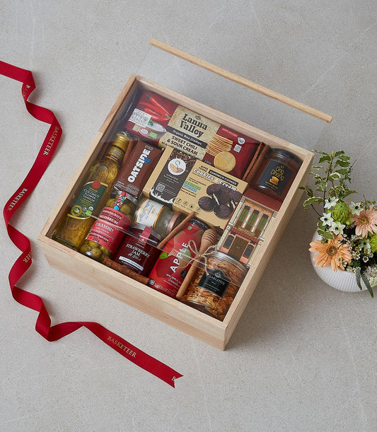 Wooden gift box filled with gourmet foods, honey, jams, and crackers with fresh flowers and red Basketeer ribbon
