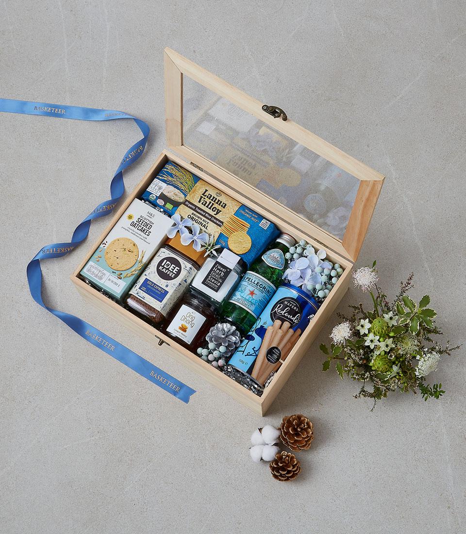 Wooden gourmet gift box with oatcakes, coffee, honey, and sparkling water, tied with blue ribbon