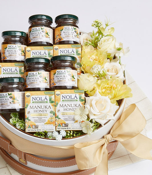 Premium Manuka honey gift basket with glass jars, yellow-orange labels, white and cream artificial flowers, and golden satin