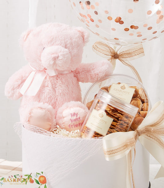 Soft pink teddy bear with gourmet cookies and crispy snacks in white gift basket with rose gold confetti balloon, perfect