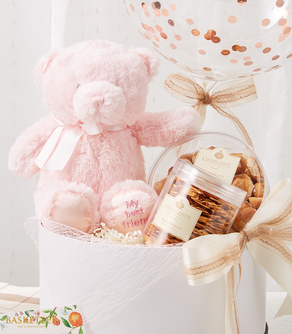 Soft pink teddy bear with gourmet cookies and crispy snacks in white gift basket with rose gold confetti balloon, perfect