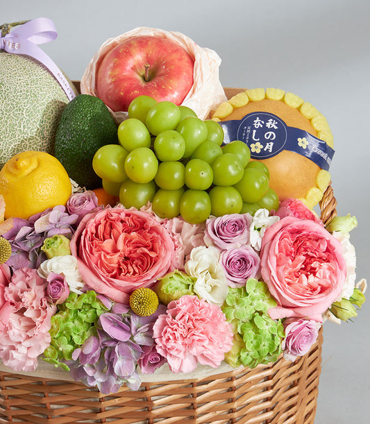 Pastel fruit and floral gift basket with fresh roses, carnations, hydrangeas, melon, grapes, and stone fruits in woven wicker