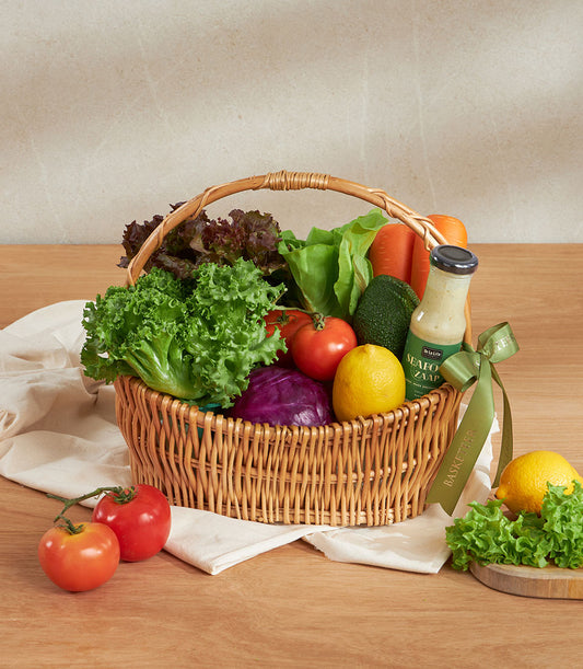 Wicker basket filled with fresh vegetables, leafy greens, carrots, tomatoes, avocado, purple cabbage, and gourmet dressing