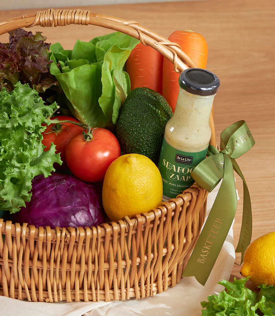 Woven wicker basket filled with fresh vegetables, lettuce, tomatoes, carrots, cabbage, avocado, lemons and creamy dressing
