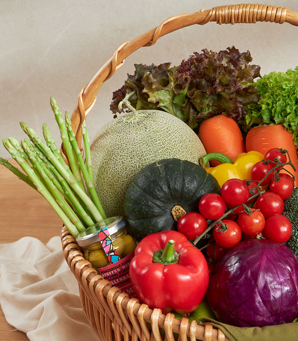 Woven wicker basket filled with fresh asparagus, cantaloupe, kabocha squash, lettuce, carrots, bell peppers, cherry