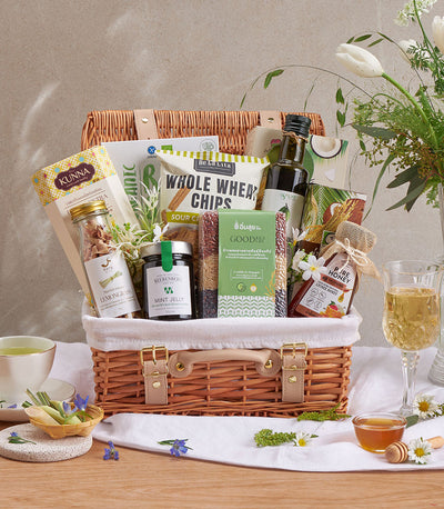 Woven picnic basket filled with gourmet wellness items including whole wheat chips, honey, lemongrass infusion, and mint