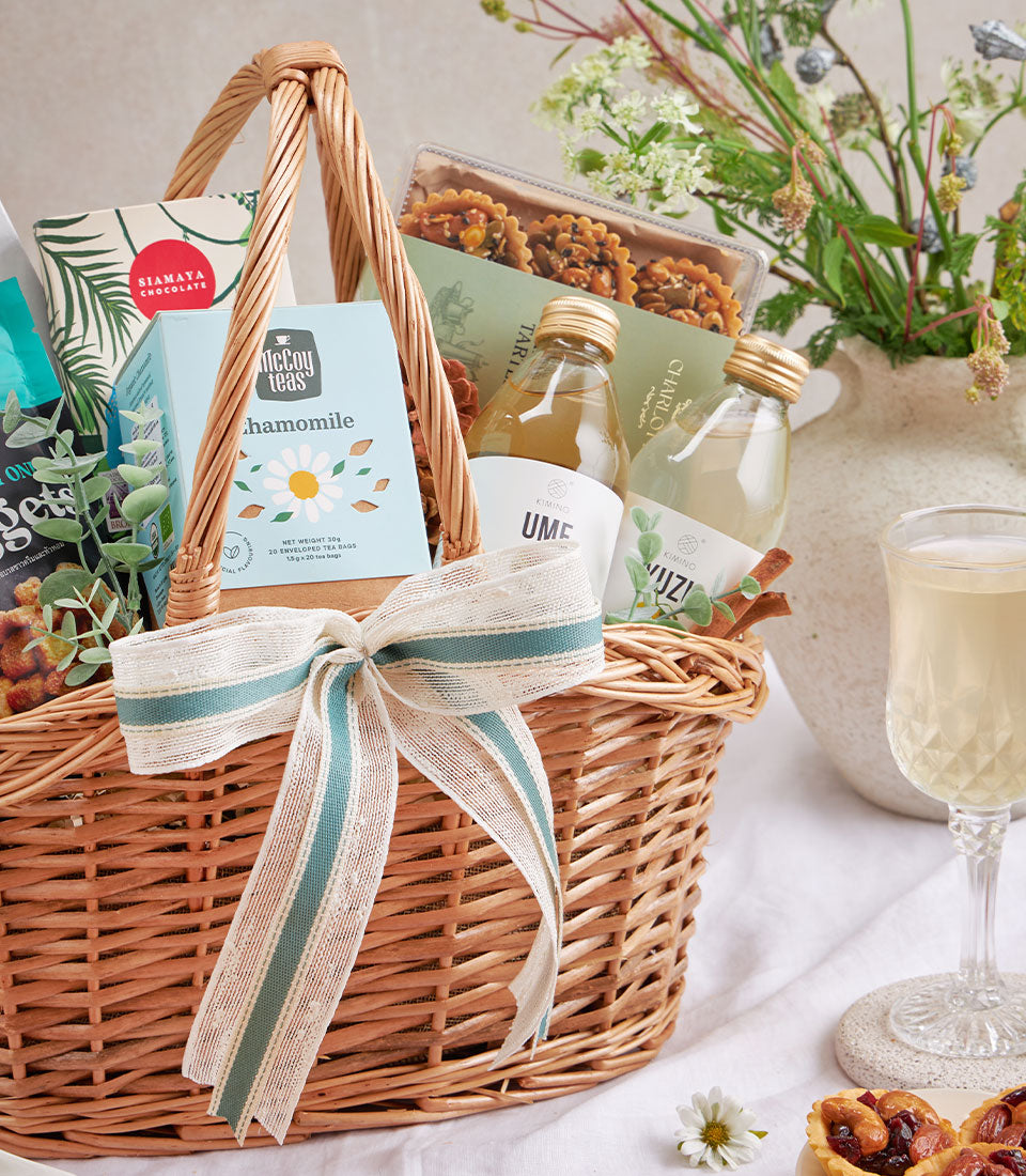 Wicker gift basket with olive oil, healthy snacks, sparkling drinks, chamomile tea, and artisan tarts tied with cream and